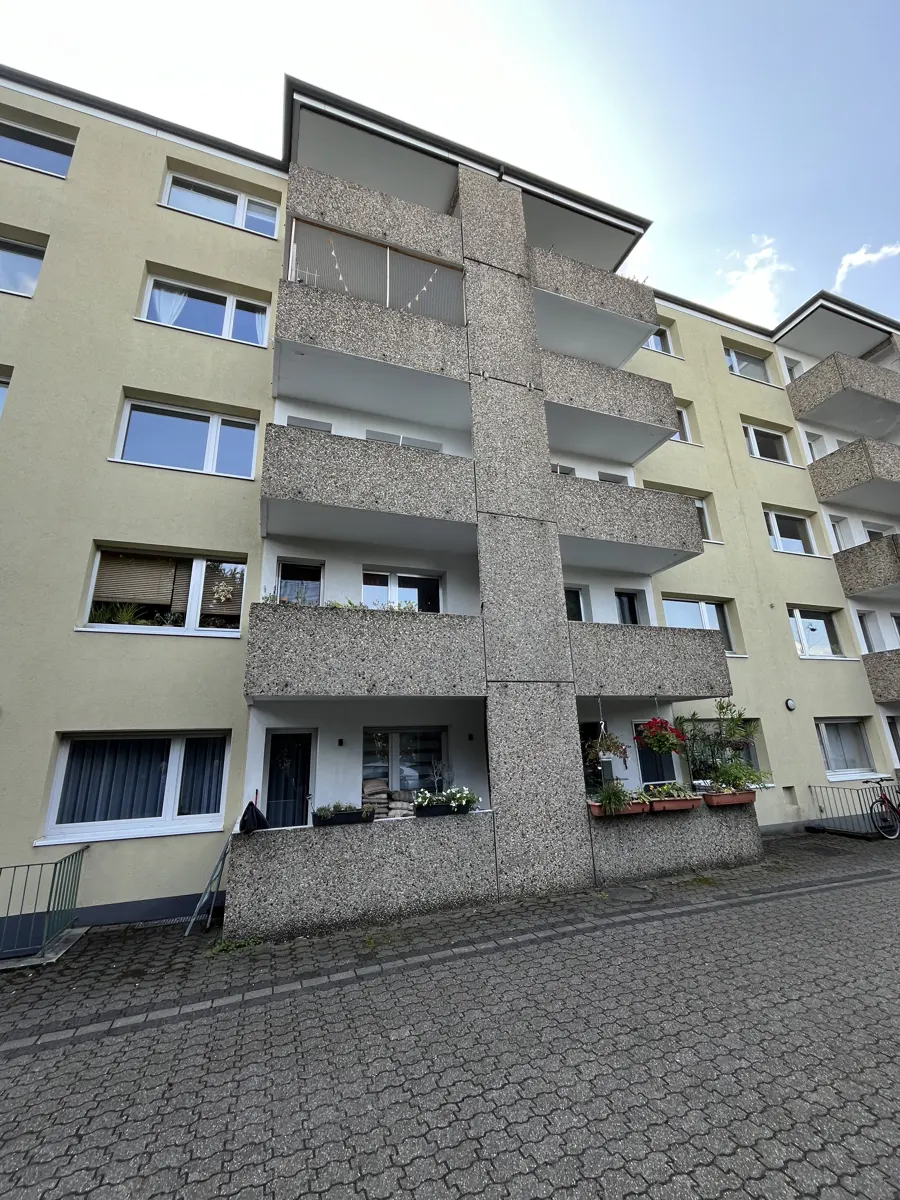 Exterior view of a multi-story apartment building with concrete balconies and a yellow facade. Windows are visible, and a paved walkway leads to the entrance.