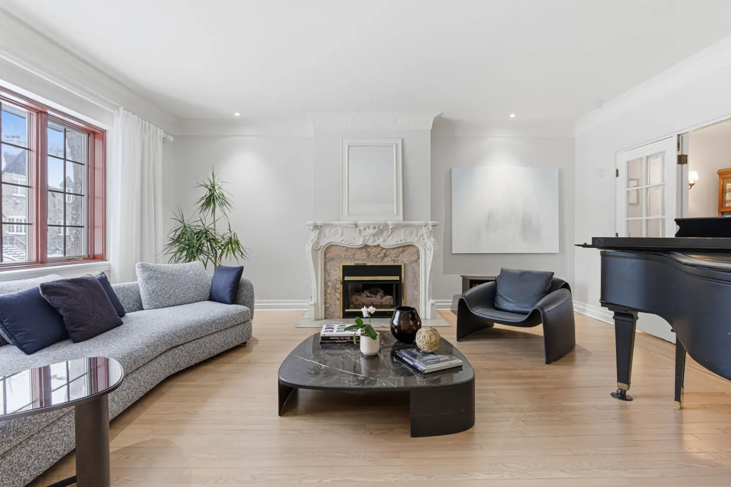 Bright living room with a gray sofa, marble fireplace, black leather chair, and a black grand piano on light wood floors.