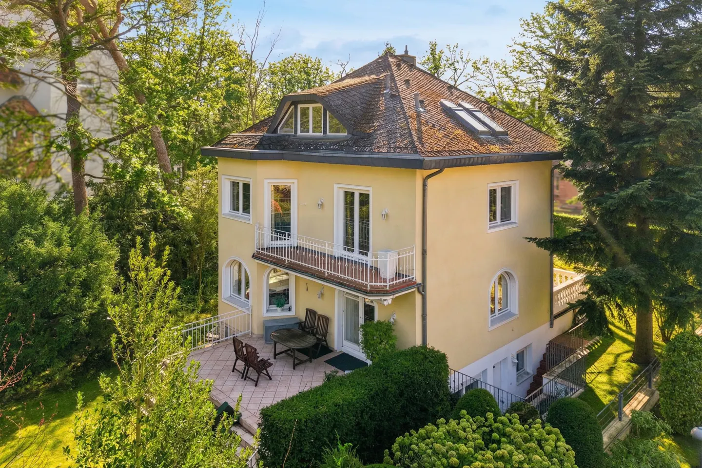 Two-story yellow house with a brown roof, white trim, and a patio with outdoor furniture, surrounded by green trees.