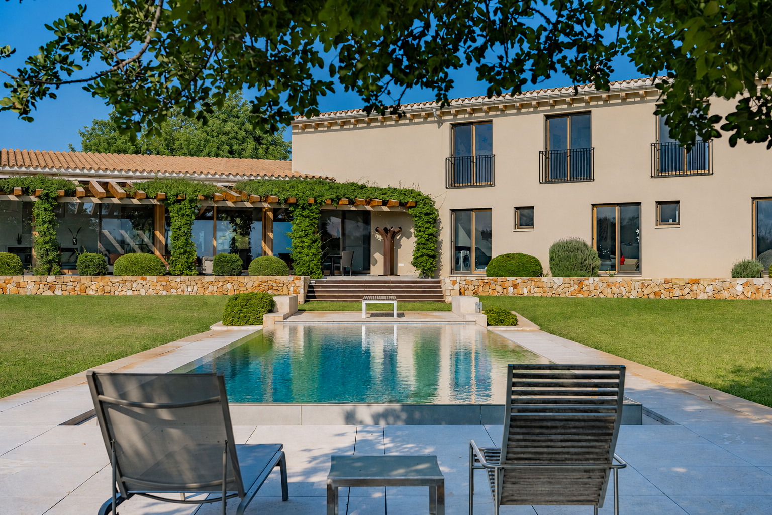 Two lounge chairs face a rectangular swimming pool in front of a beige two-story villa with a vine-covered pergola. [AI-generated text]