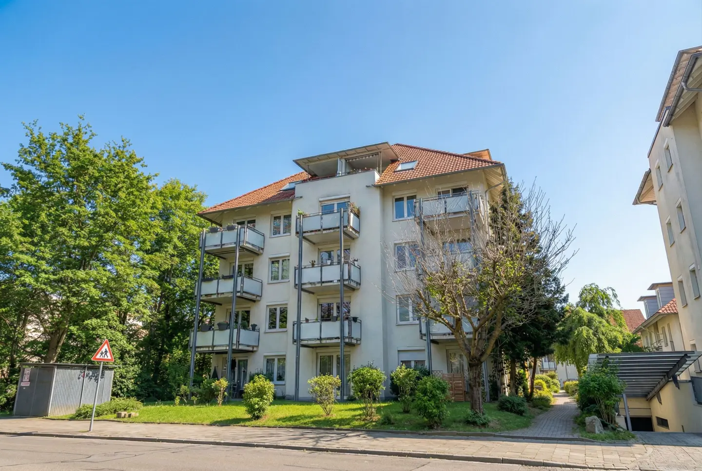 Exterior view of a light-colored apartment building with balconies, a red tile roof, and green trees under a clear blue sky.