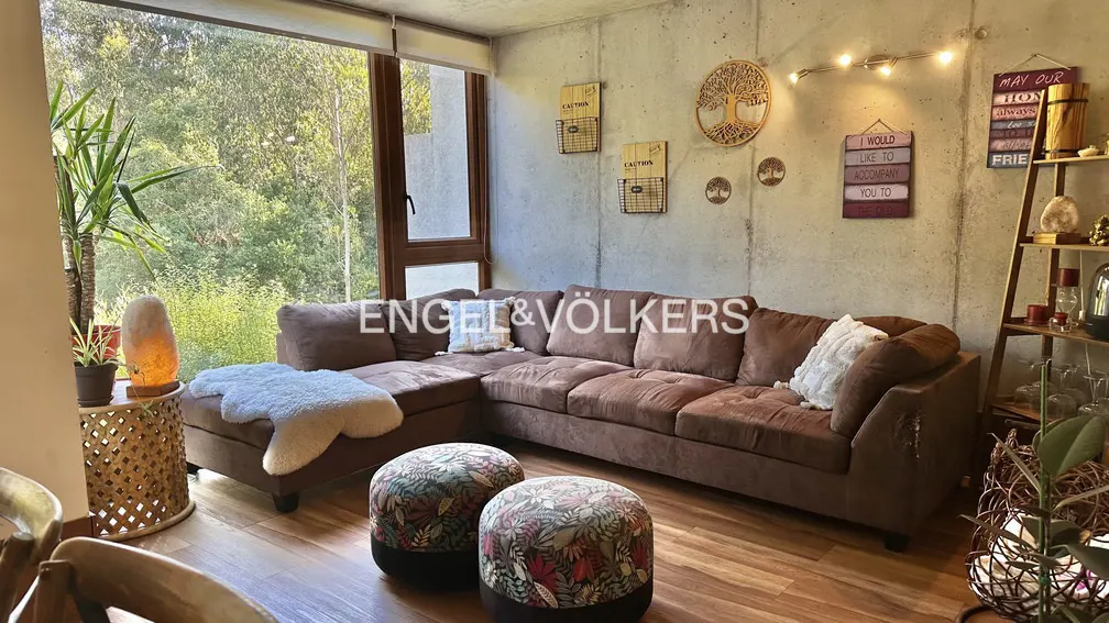 Living room with brown sectional sofa, floral ottomans, wood floors, and concrete walls decorated with wooden art. Large window with green trees outside.