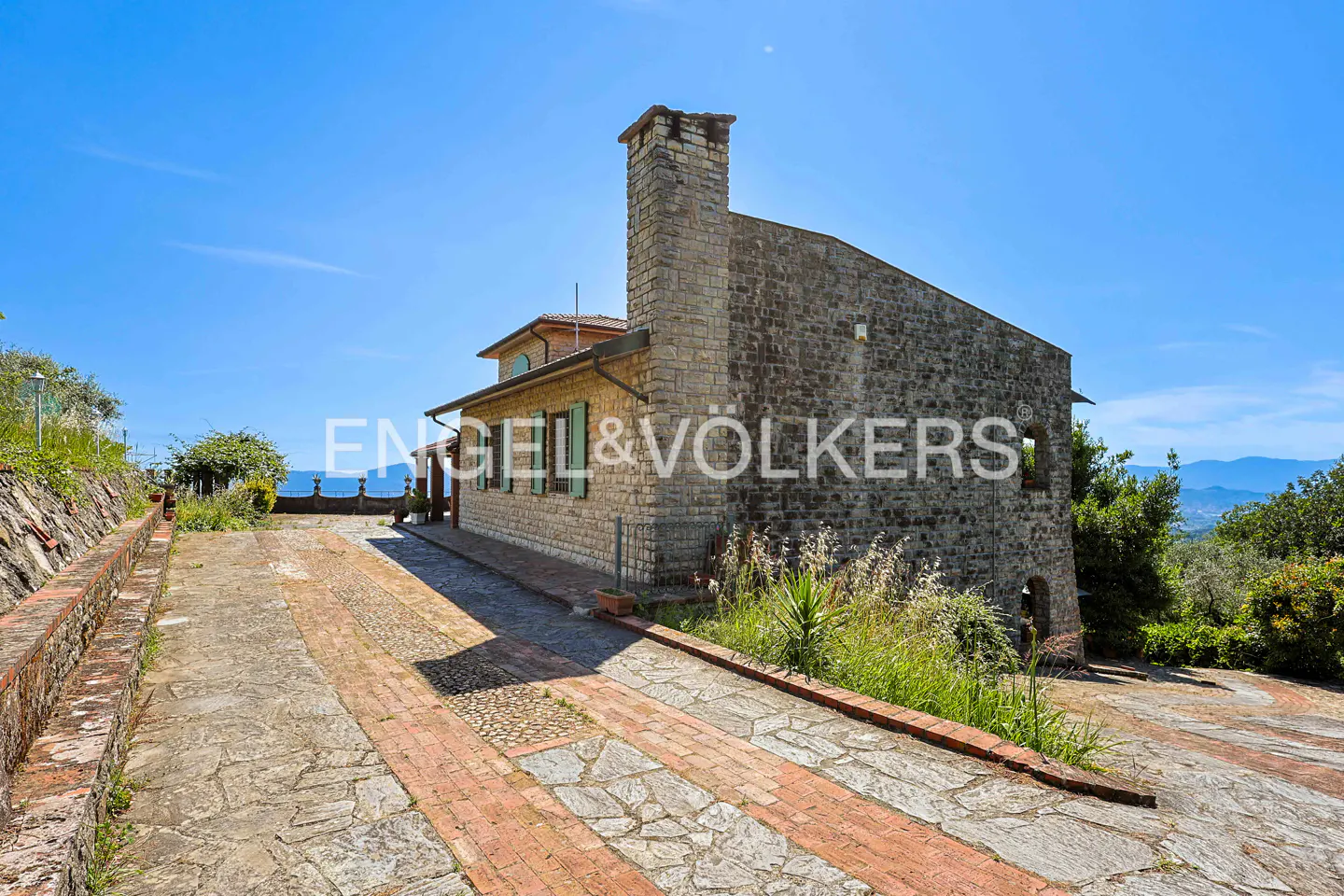 A stone house with a tall chimney sits on a paved driveway under a clear blue sky, surrounded by lush greenery.