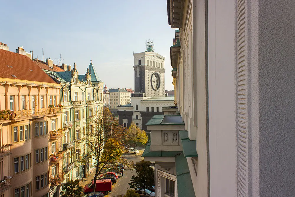 View from a balcony overlooking a European city street with historic buildings and a prominent clock tower in the distance.