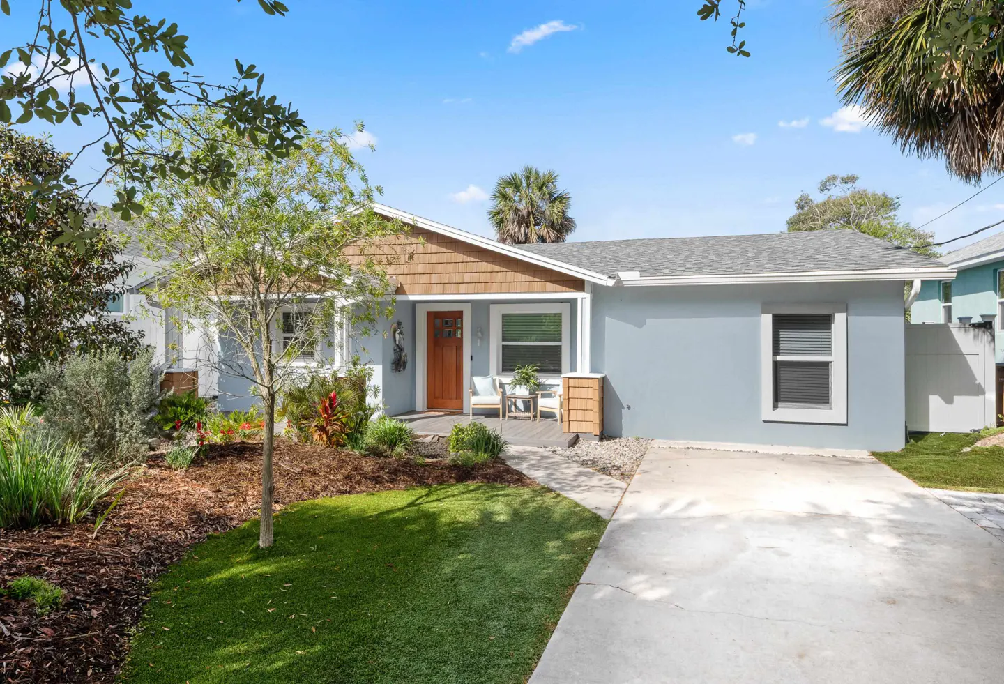 Light blue single-story home with a brown wood door, gray roof, green lawn, and concrete driveway.