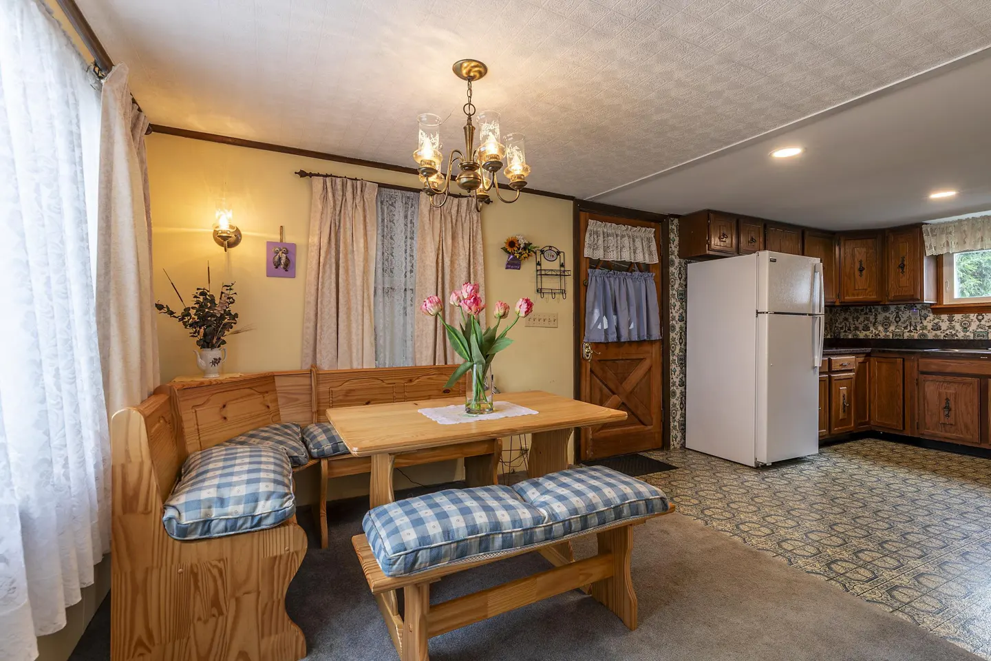A kitchen and dining area with a wooden corner booth, table, and bench with blue plaid cushions. A vase of pink tulips sits on the table under a chandelier. White fridge and wood cabinets in the background.