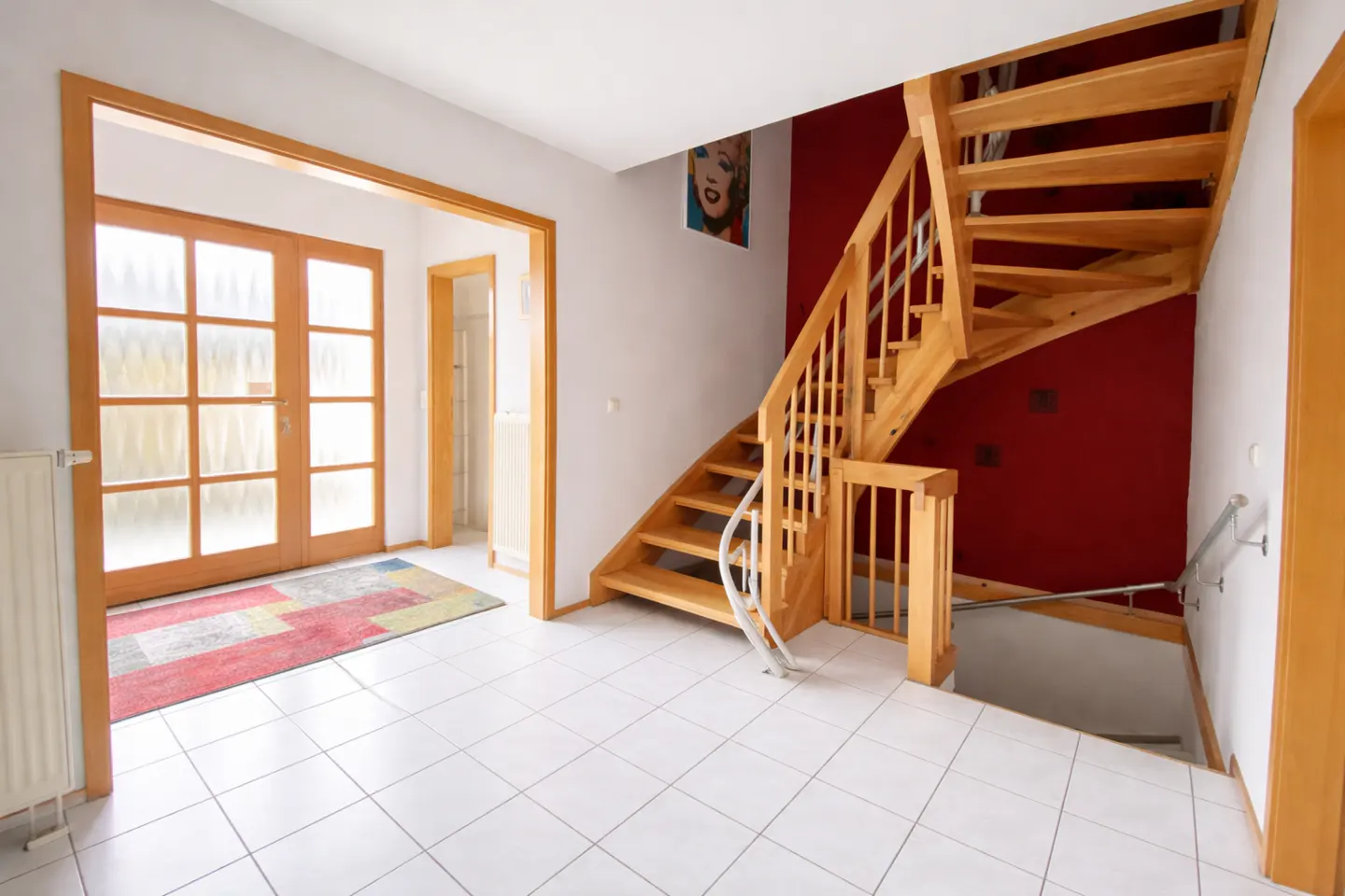 Bright foyer with white tile floor, wooden staircase against a red wall, and glass paneled doors leading outside. A colorful rug sits by the entrance.