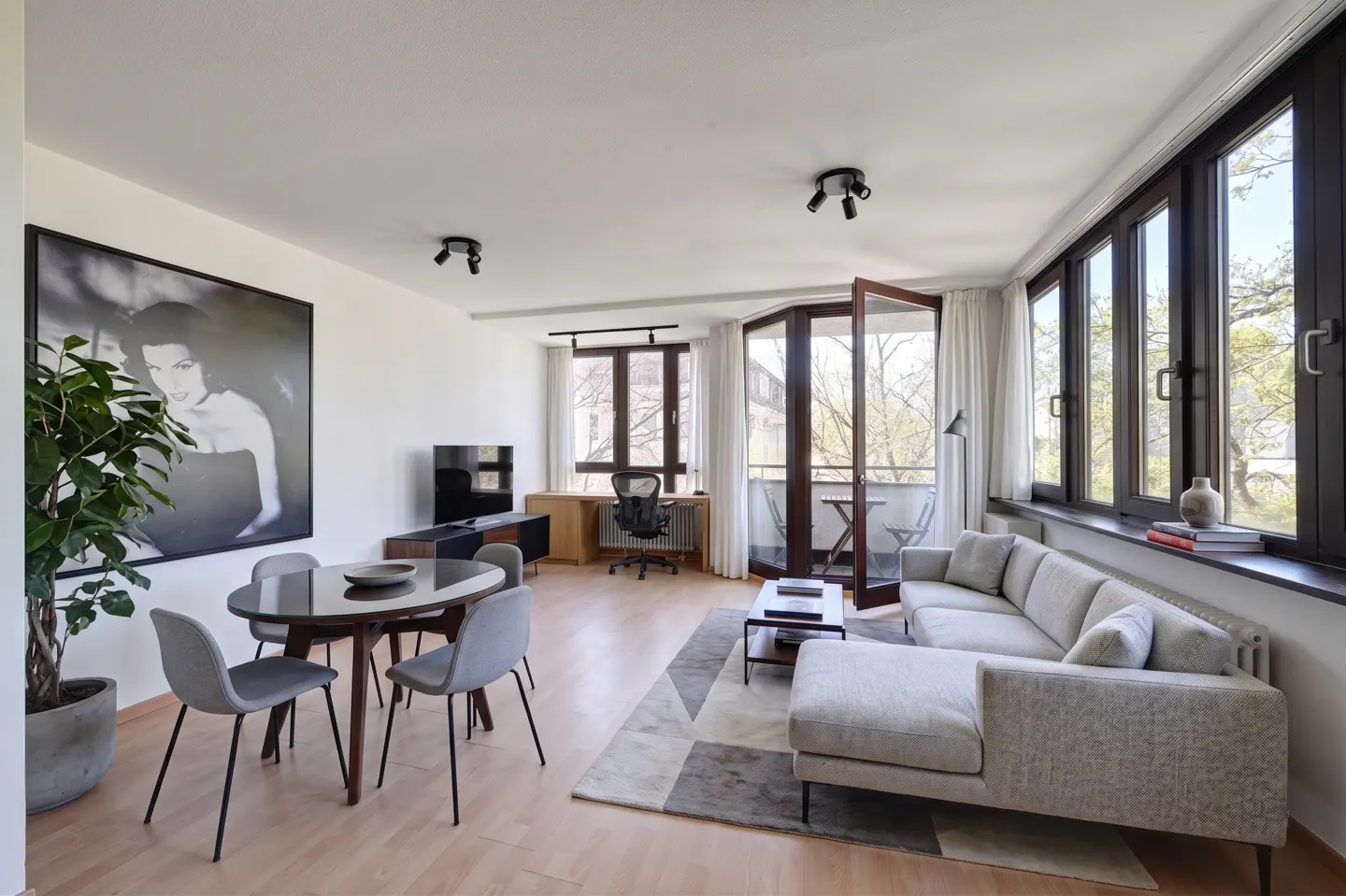 Bright living room with wood floors, a gray sectional sofa, a round table with gray chairs, and a balcony with open doors.