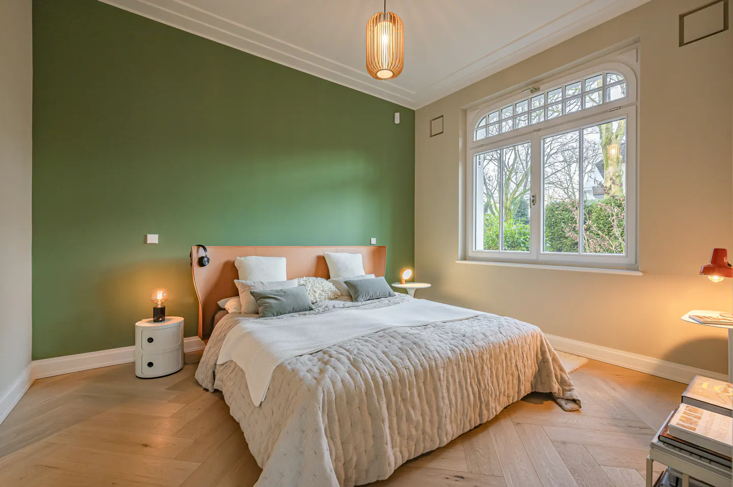 Bedroom with green wall, wood floors, and a large window. A bed with a tan headboard and a light quilt is centered. Nightstands with lamps flank the bed.