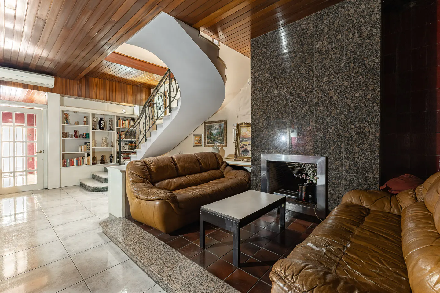 Living room with two brown leather sofas, a coffee table, a granite fireplace, and a curved staircase with wood ceiling.