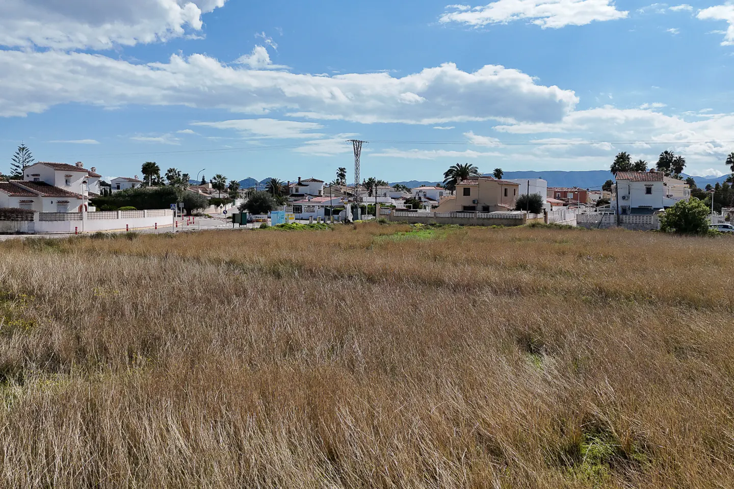 Vacant lot with dry grass in a residential area under a blue, cloudy sky. Houses and power lines are visible in the background.