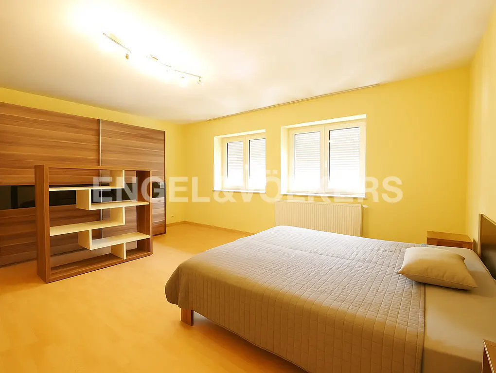 Bedroom with a gray bed, yellow walls, and wood closet. Windows provide natural light. A bookshelf stands near the closet.