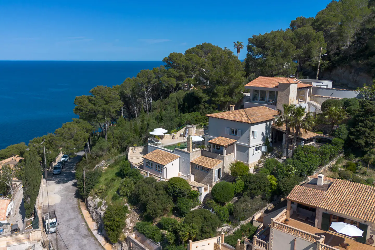 Aerial view of a multi-level home with a red tile roof, surrounded by green trees, overlooking the blue ocean.