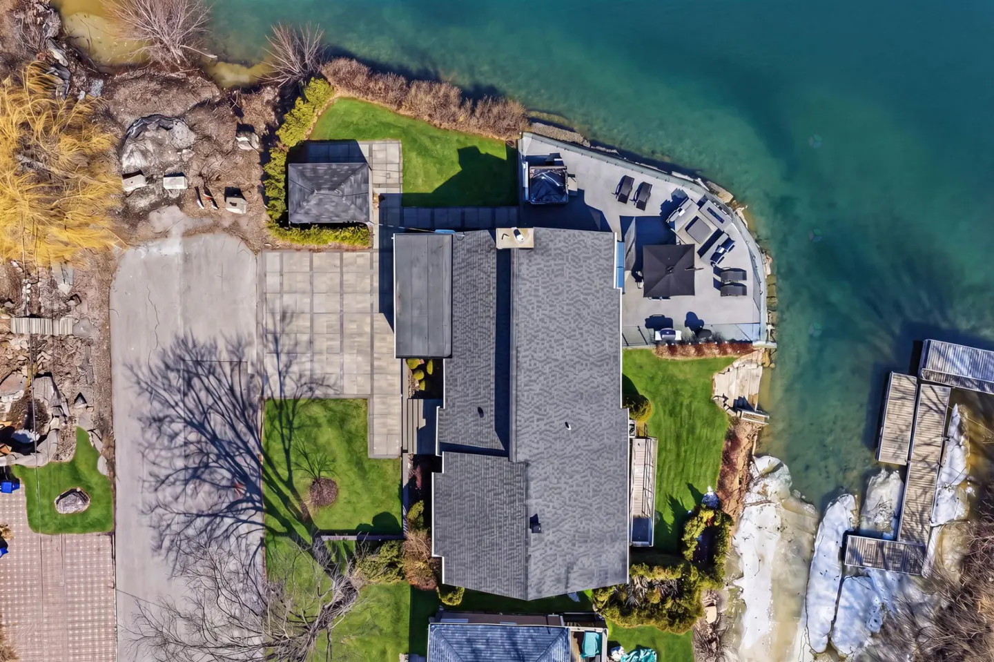 Aerial view of a large house with a dark roof, a patio with lounge chairs, and a dock on a turquoise lake.