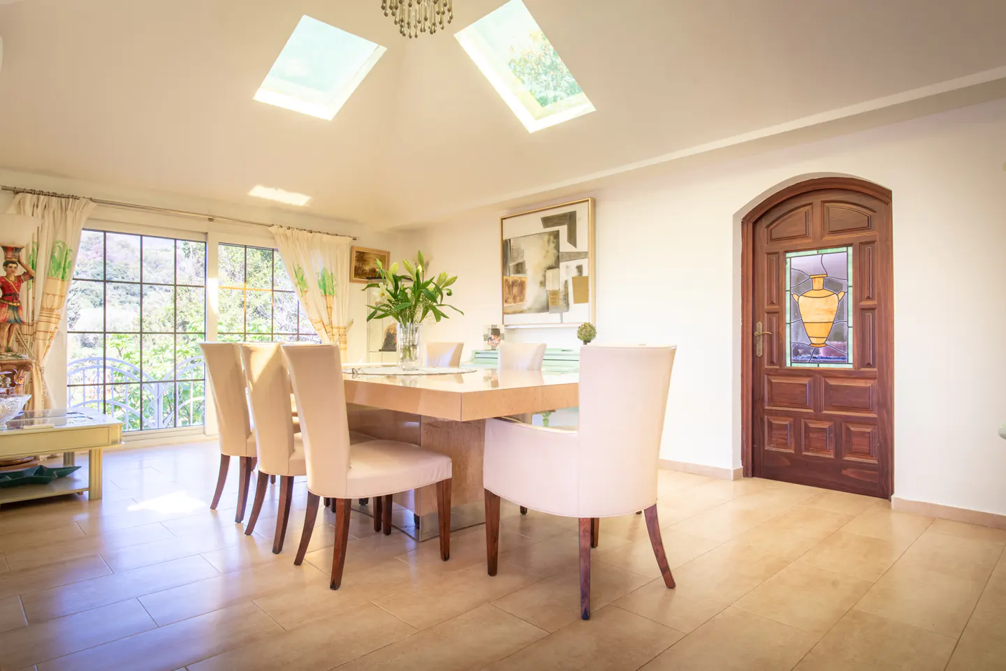 Bright dining room with a light wood table, cream chairs, and a stained-glass door. Skylights and a large window provide natural light.