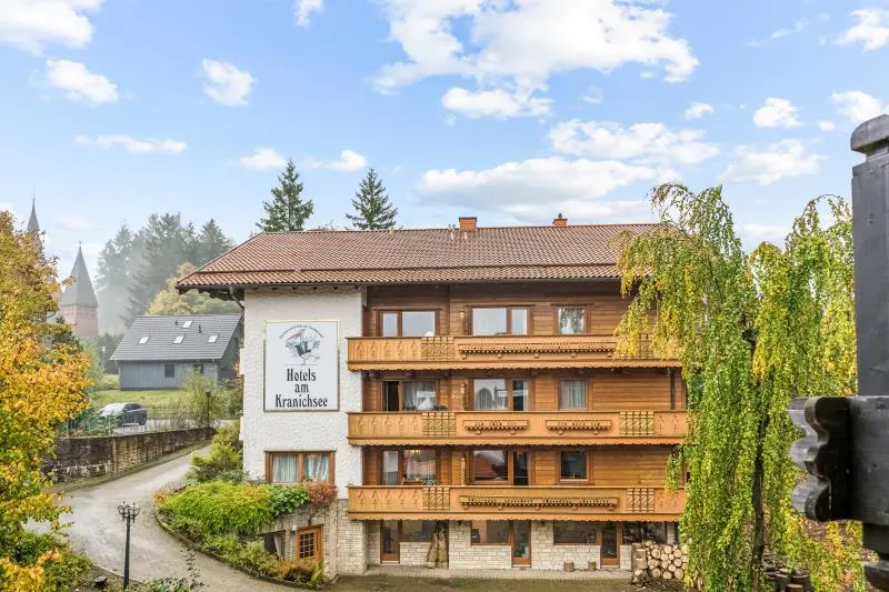 Exterior view of Hotels am Kranichsee, a multi-story building with wood balconies, set against a cloudy sky and trees.