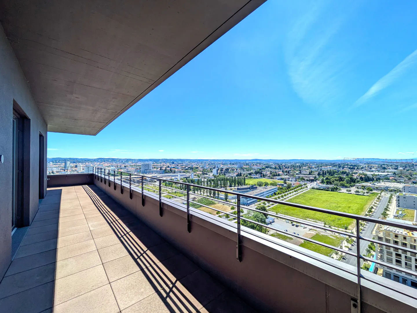 Spacious high-rise balcony with tiled flooring and metal railings overlooking a sunny city landscape under a clear blue sky.