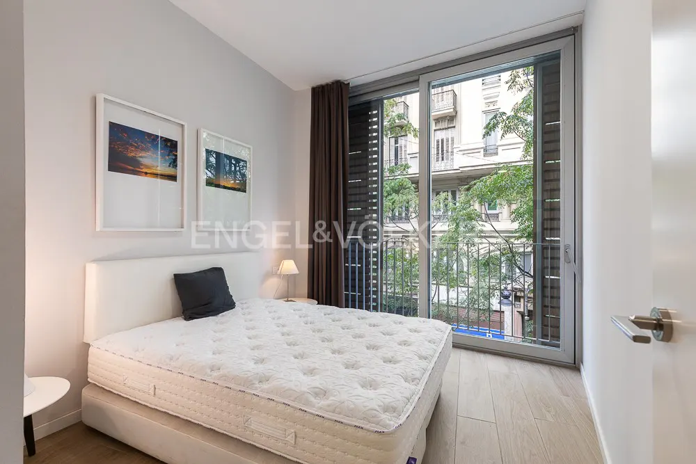 Bedroom with white bed, gray pillow, and balcony view of a European city street with trees. Two framed pictures on the wall.