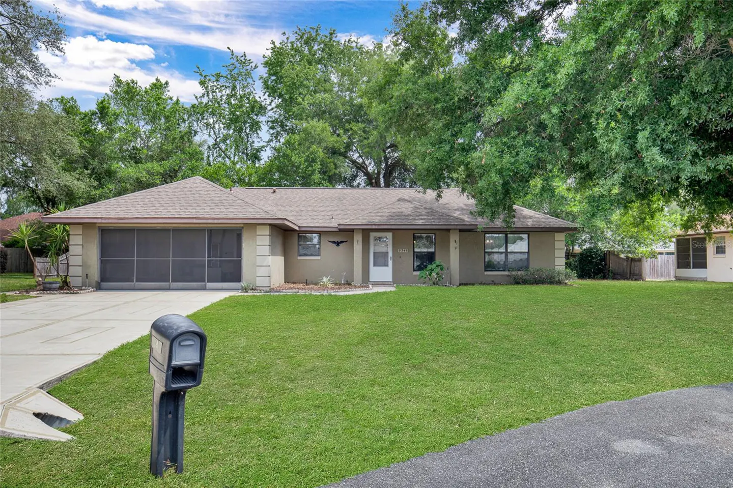 Single-story beige house with a brown roof, green lawn, and trees in the background. A black mailbox stands in the front yard.