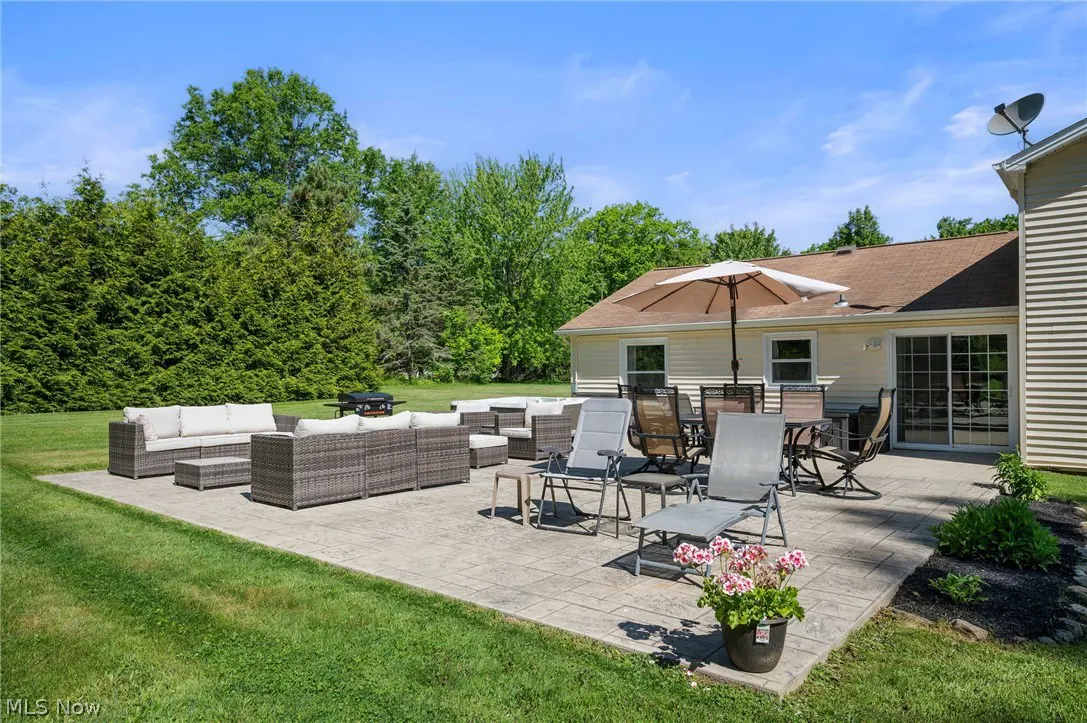 Spacious backyard patio with gray wicker sectional seating, dining set with umbrella, lounge chairs, and lush green trees.