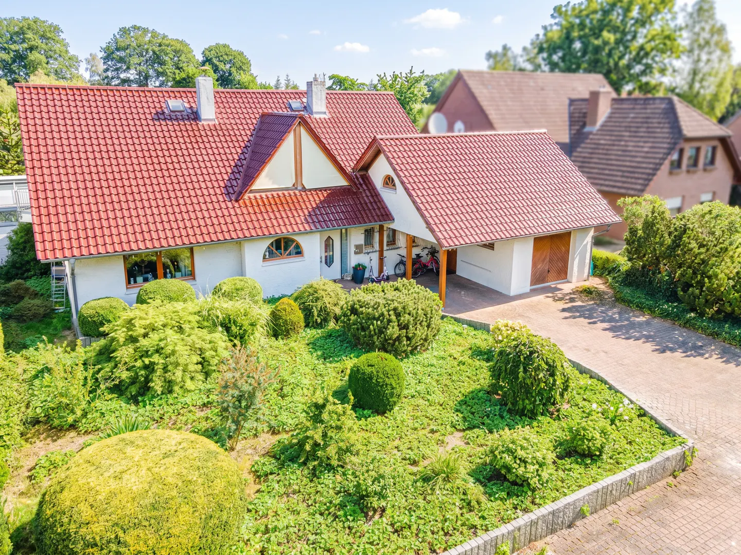 White house with a red tiled roof, a carport, and a lush, well-manicured garden with rounded shrubs and a paved driveway.
