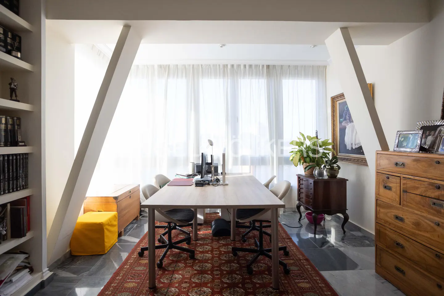Bright office space with a long table, chairs, and a red patterned rug. Bookshelves and wooden furniture add character to the room.