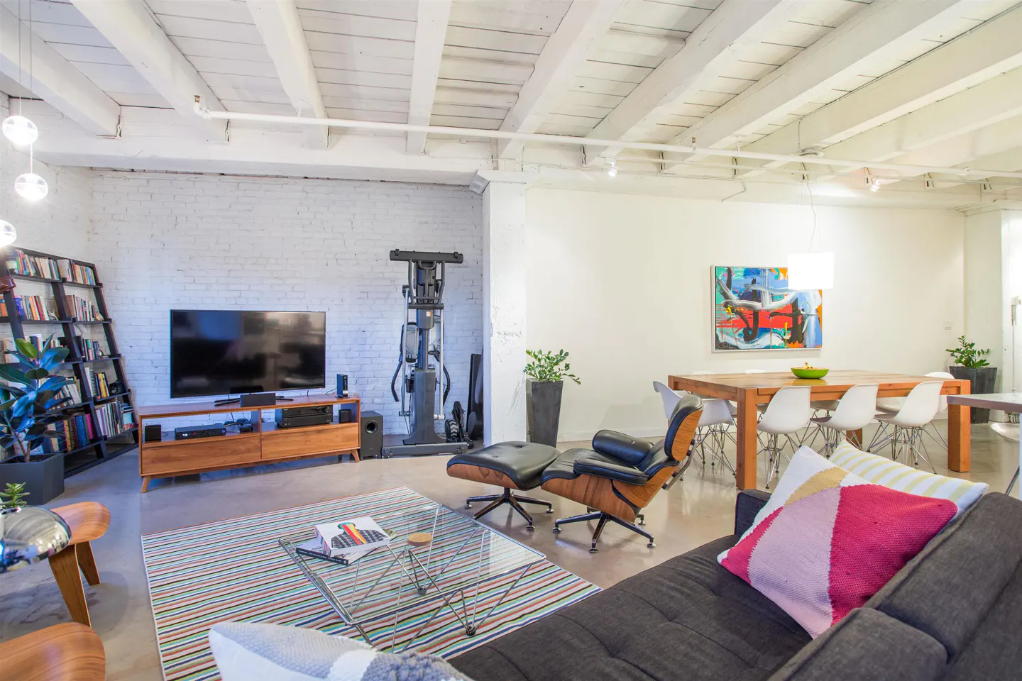 Bright, open living space with white brick walls, exposed beams, and a mix of modern furniture. A large TV, dining table, and exercise equipment are visible.