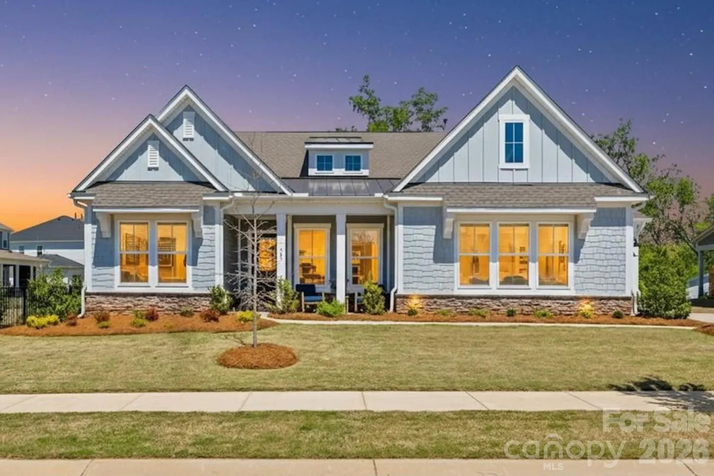 Light blue craftsman-style home with stone accents, glowing windows, and a sunset sky at dusk.