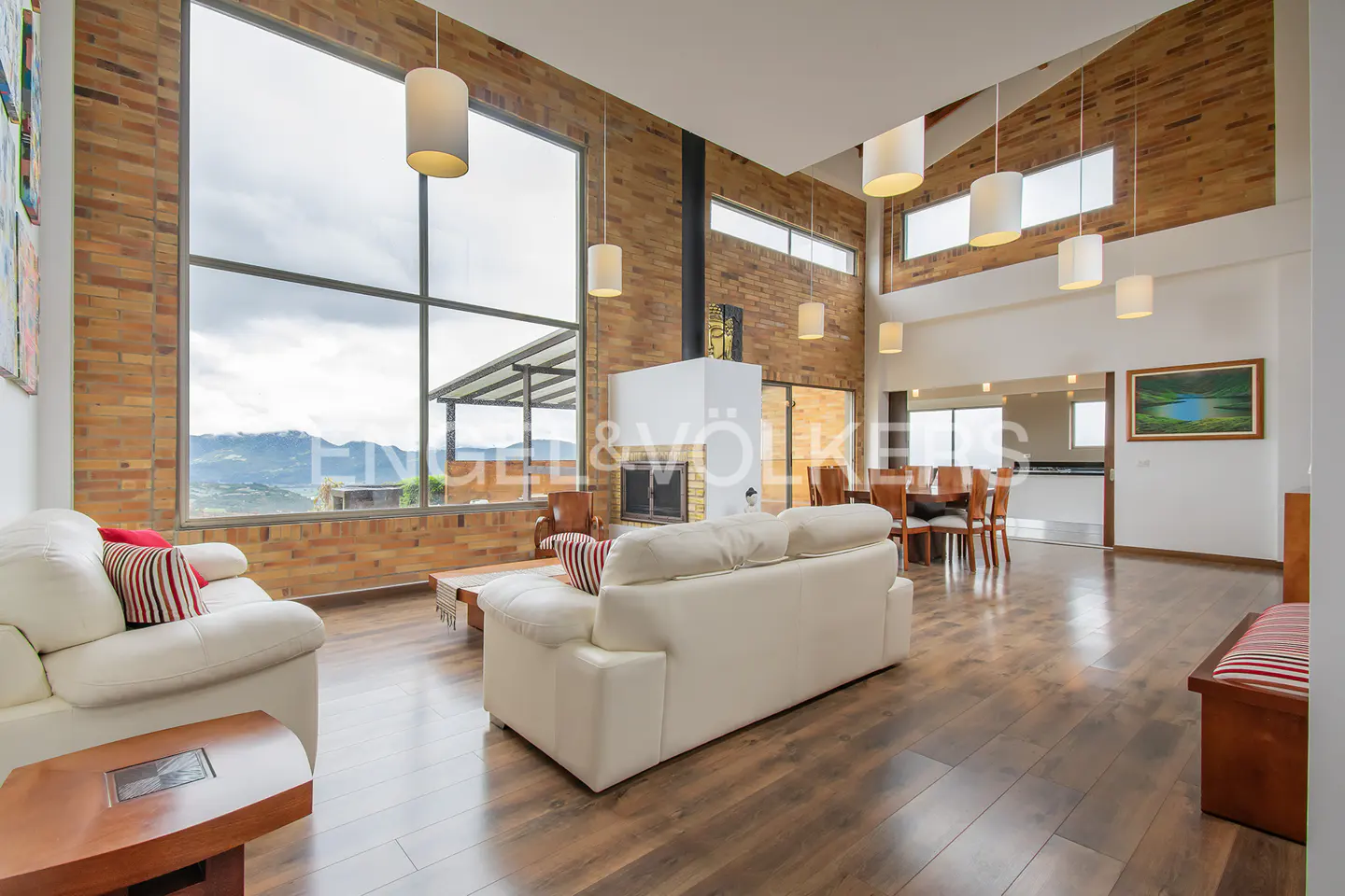 Bright living room with brick walls, wood floors, and white sofas. Large windows offer mountain views. Dining area visible in the background.