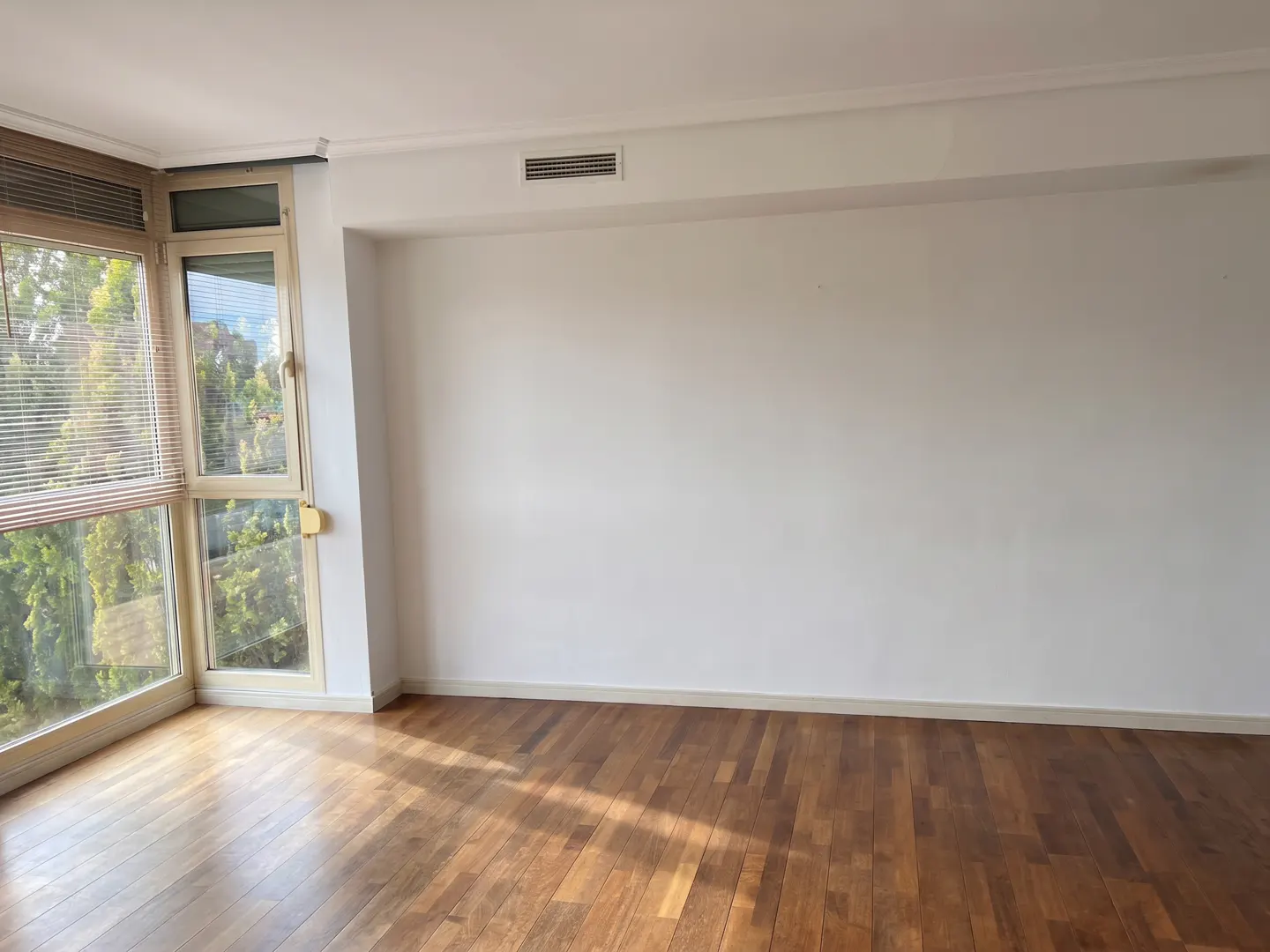 Empty room with white walls, dark hardwood floors, and large windows looking out onto green trees.