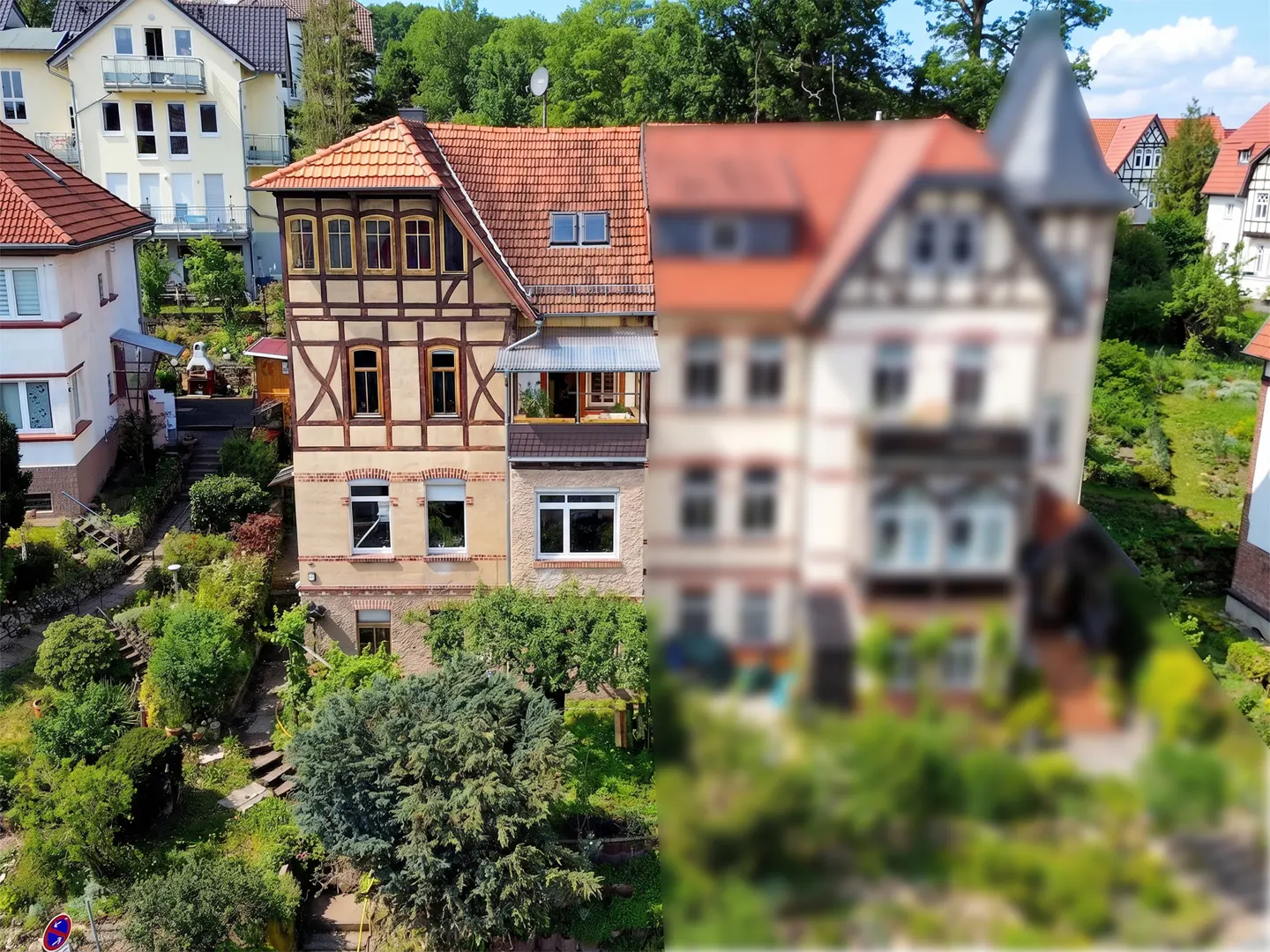A half-timbered house with a red tile roof stands among lush greenery. Another blurred house is on the right.