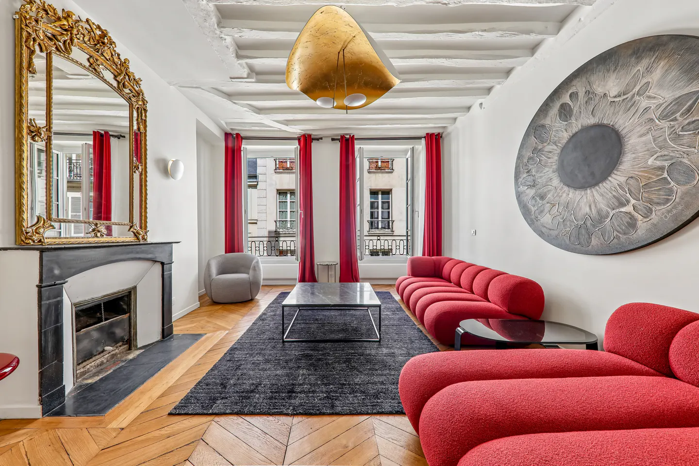 Living room with red sofa, black rug, and fireplace. Windows with red curtains overlook city buildings. A large eye art piece hangs on the wall.