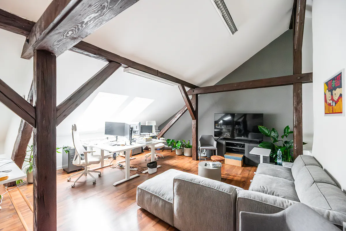 Attic office space with exposed dark wood beams, white walls, and hardwood floors. A gray sofa and white desks with computers furnish the room.