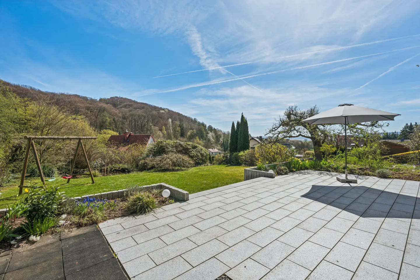 A backyard patio with gray pavers, a green lawn, a swing set, and a gray umbrella under a blue sky.