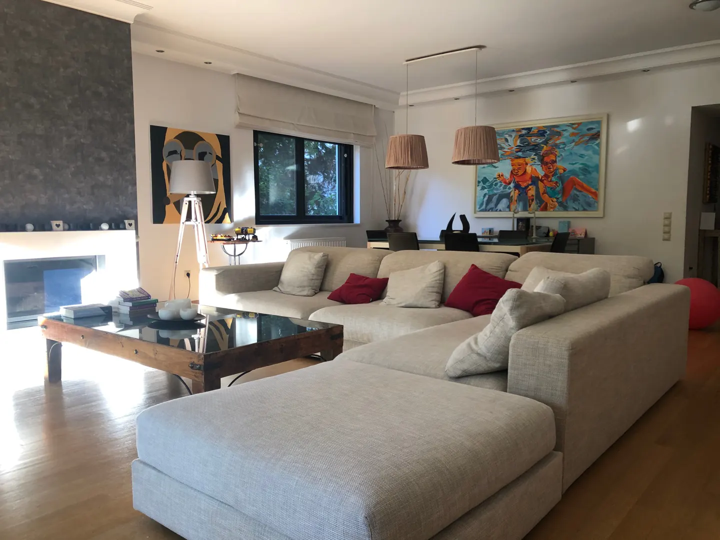 Bright living room with a large beige sectional sofa, wood floor, and modern art. A fireplace and coffee table are also visible.