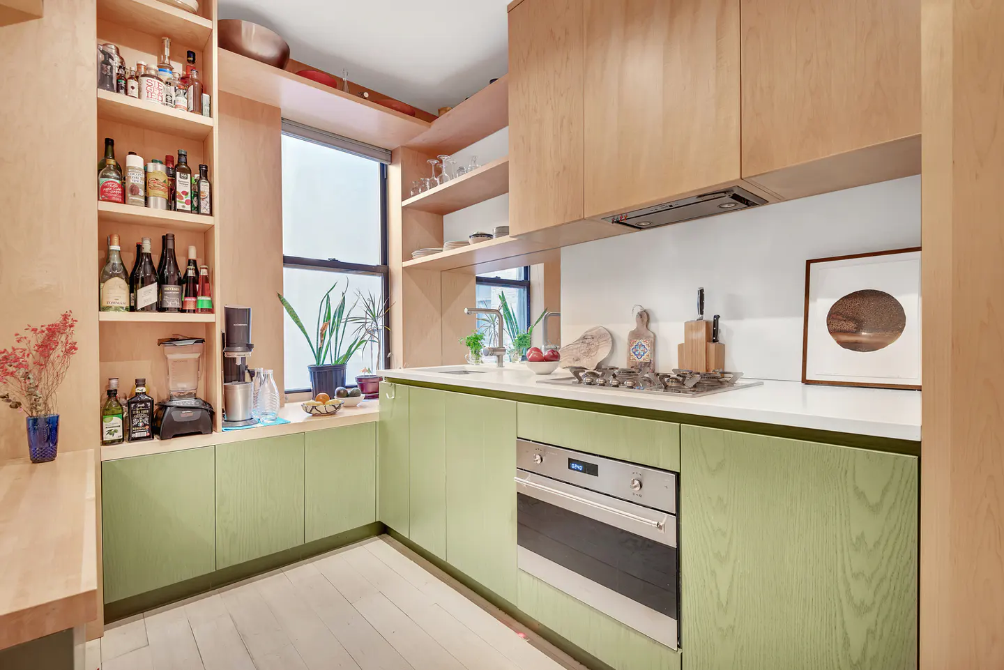 A modern kitchen with light green cabinets, white countertops, and light wood shelving. A window is above the sink.