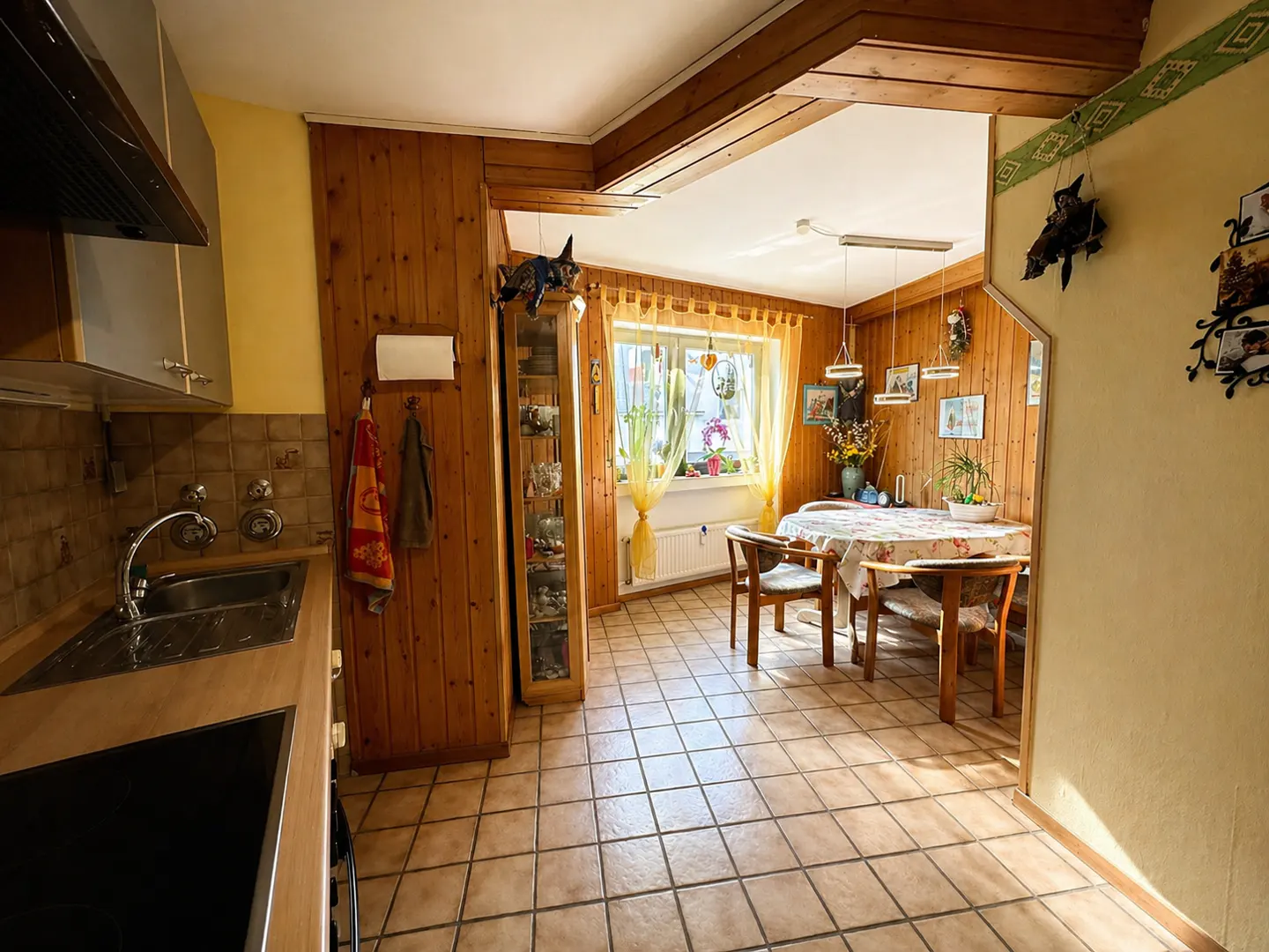 A bright kitchen and dining area with wood paneling and tile floors. A table with chairs sits near a window with yellow curtains.