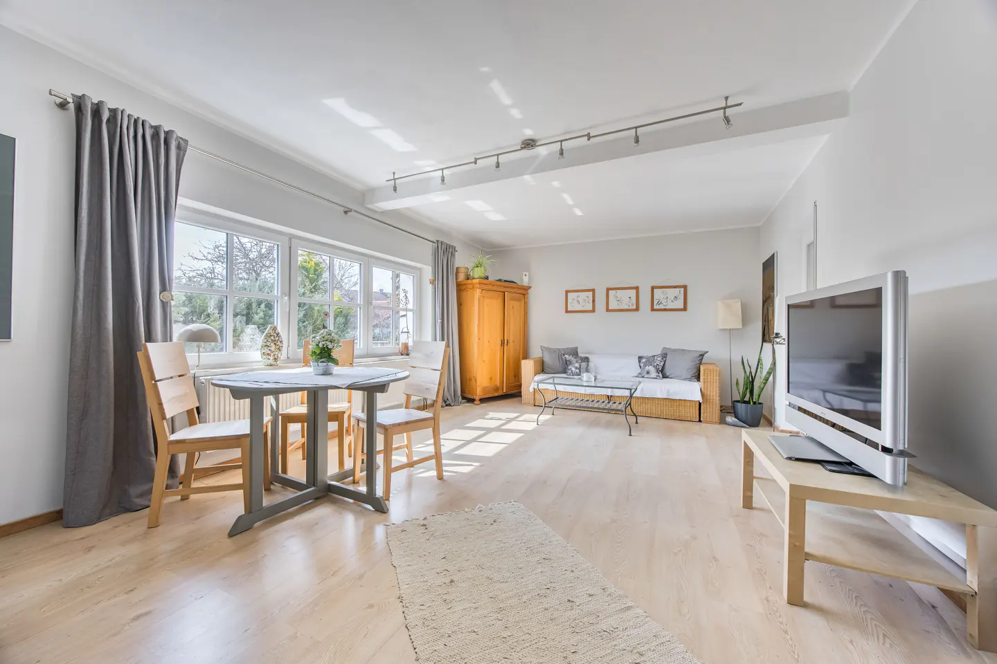 Bright living room with light wood floors, a gray table and chairs, a wicker couch, and a large television.