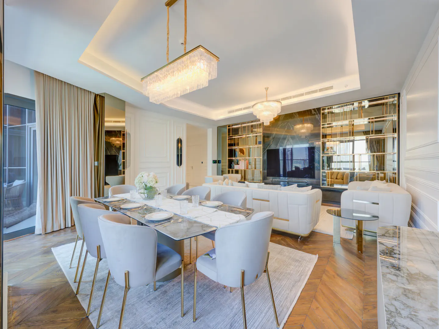 A bright, modern dining and living room with a long table, gray chairs, and a crystal chandelier. White sofas and a mirrored wall are in the background.