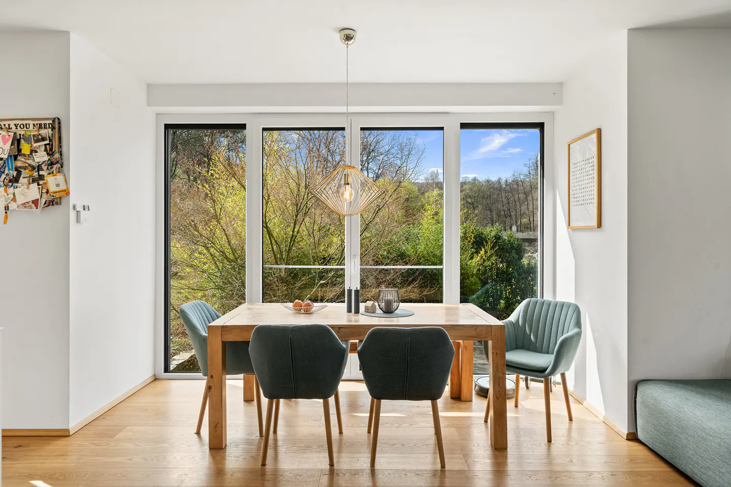 Bright dining room with a wood table, four chairs, and a modern light fixture. Large windows overlook a green, wooded area.