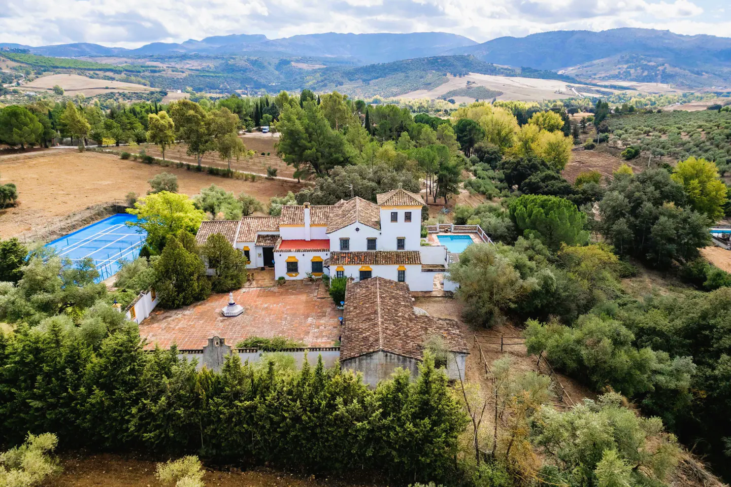 Aerial view of a white Spanish-style villa with a red tile roof, pool, tennis court, and green trees against a mountain backdrop.