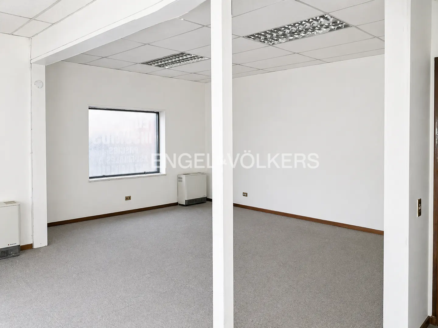 Bright, empty office space with gray carpet, white walls, and a window. A white support beam divides the view. Engel & Völkers logo on the wall.