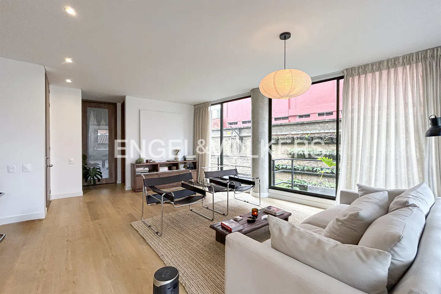 Bright living room with wood floors, beige sofa, two black chairs, and large windows with sheer curtains.