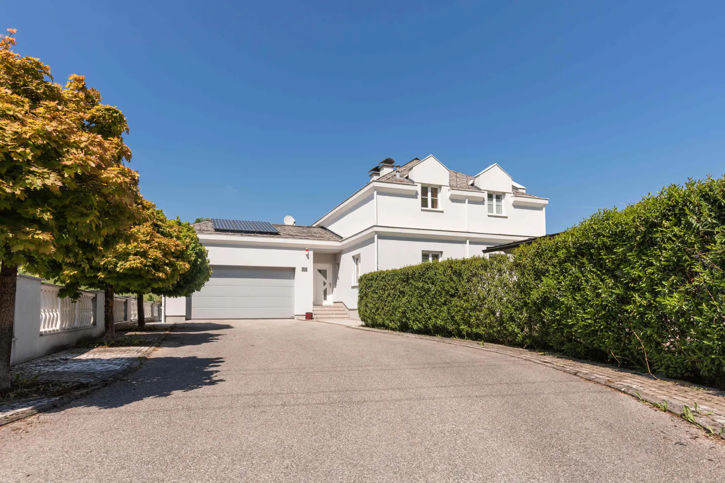 Exterior of a two-story white house with a gray garage door, solar panels, and a green hedge.