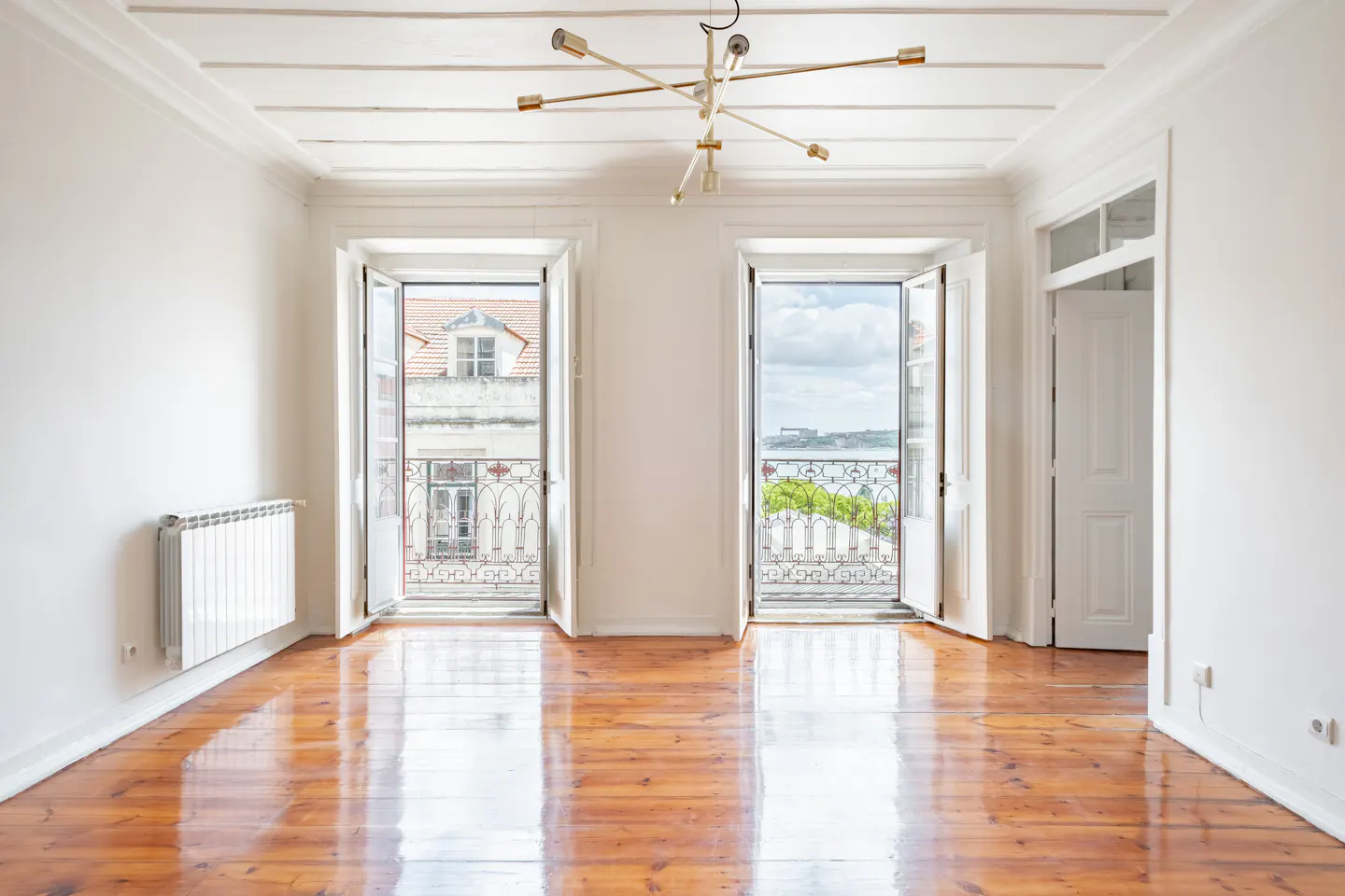 Bright, empty room with shiny wood floors, white walls, and two open balcony doors with city views. A modern chandelier hangs from the ceiling.