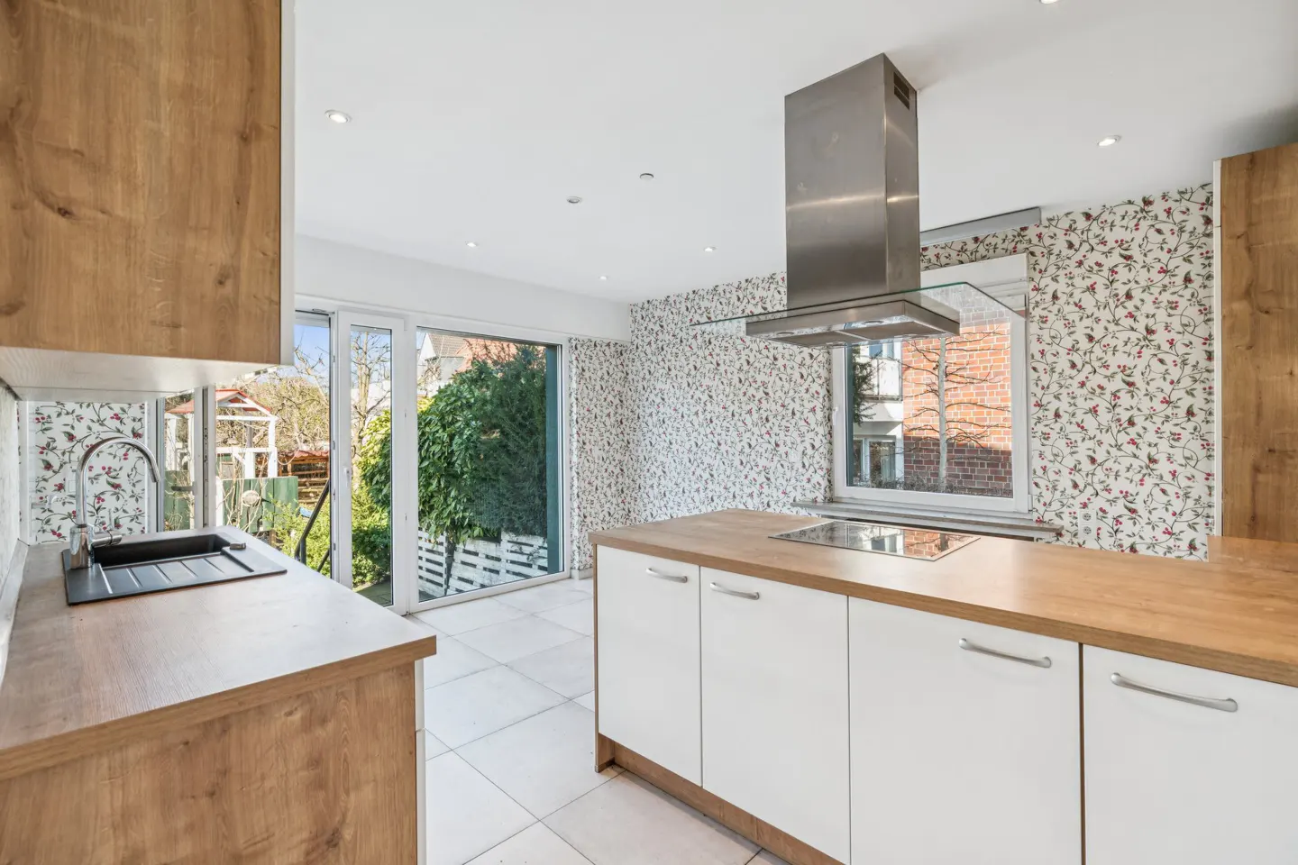 Bright kitchen with white cabinets, wood countertops, and floral wallpaper. A stainless steel range hood hangs above the cooktop. A glass door leads to a garden.