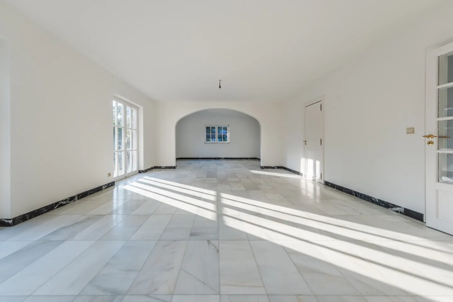 Bright, empty room with white walls, marble floors, and black trim. Sunlight streams through windows and an arched doorway.