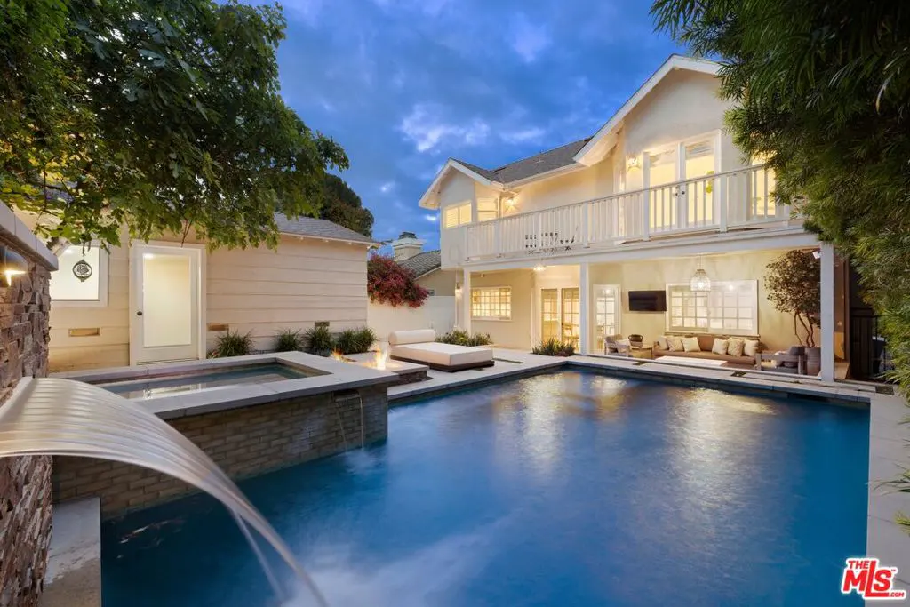 Backyard view of a two-story house with a pool, spa, and outdoor seating area. The pool has a waterfall feature and is surrounded by trees.