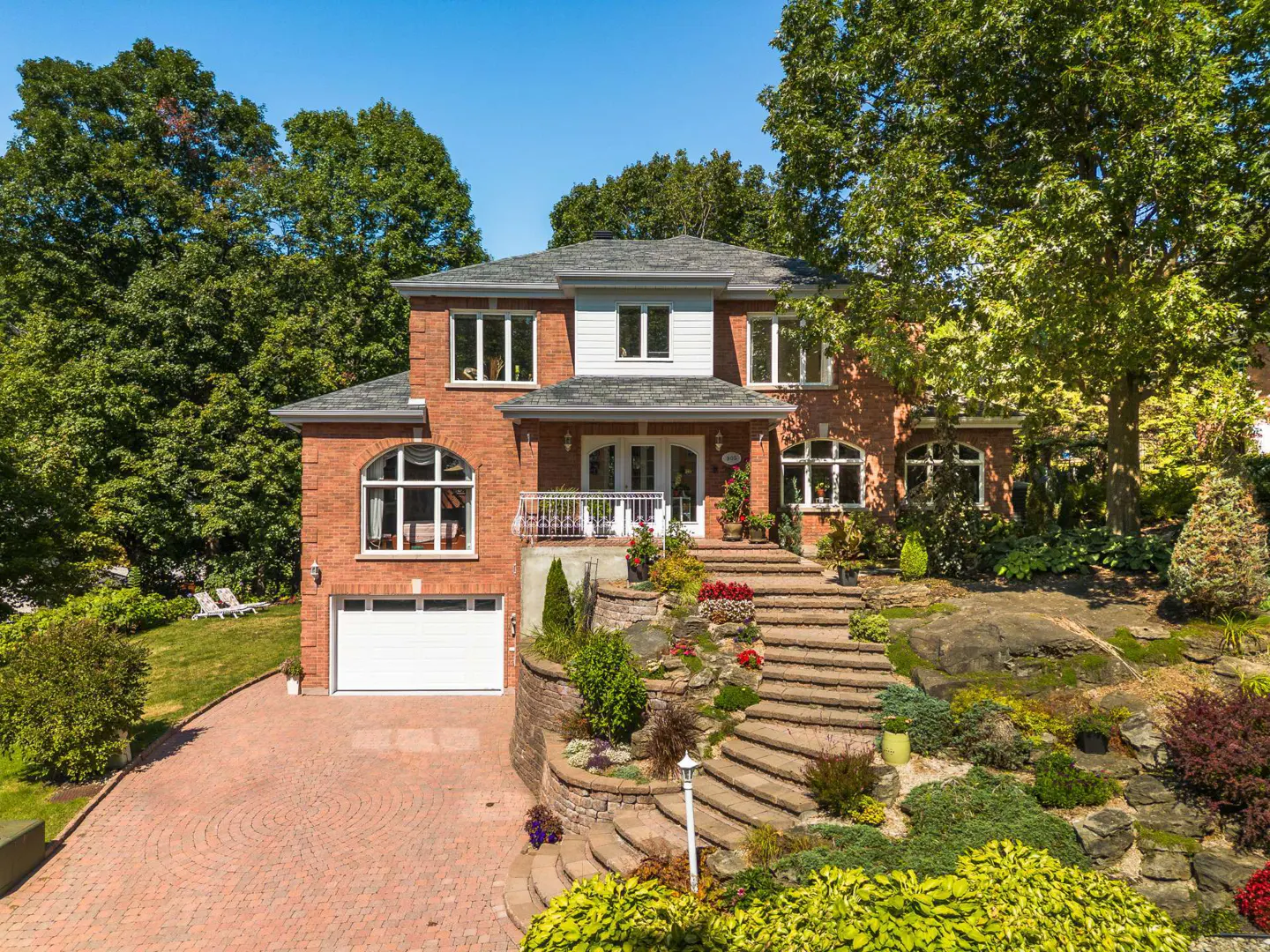 Brick house with white trim, gray roof, and a white garage door. Stone steps lead to the front door, surrounded by lush greenery.