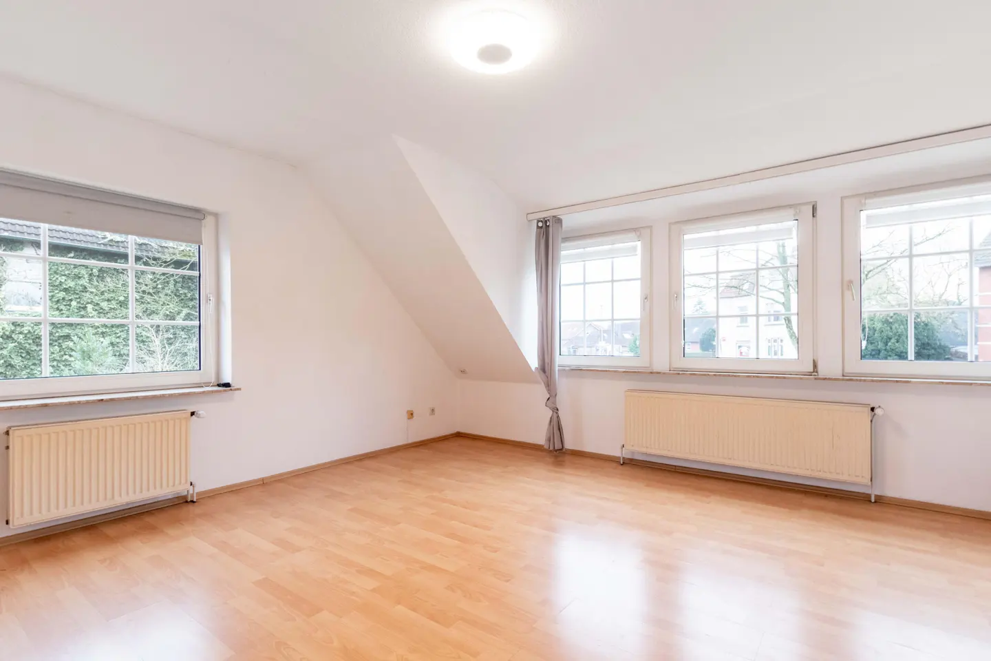Bright, empty room with wood floors, white walls, and multiple windows. Radiators are under the windows.